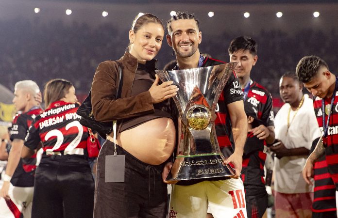 Arrascaeta e sua esposa Camila, grávida do primeiro filho do casal, com a taça de Campeão Brasileiro 2025 - Foto: Adriano Fontes / Flamengo